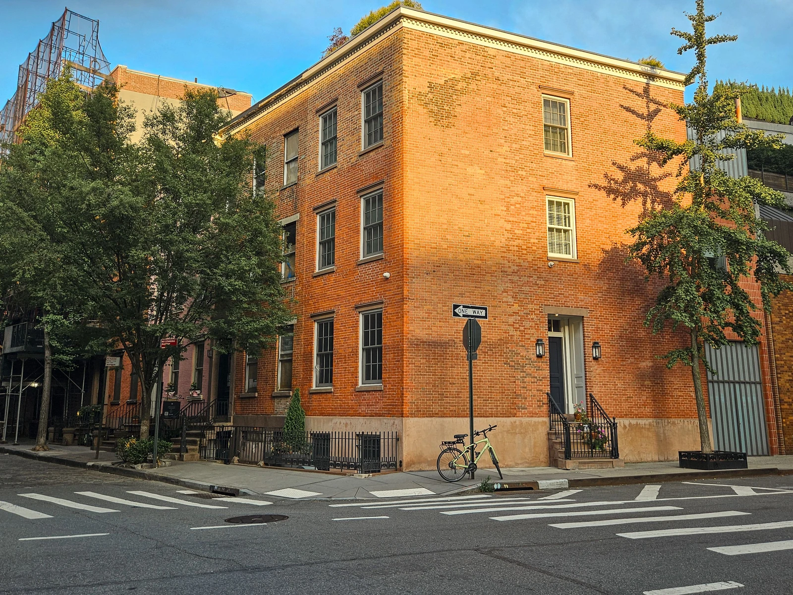Golden sunlight across a red-brick corner house in New York, with dark-framed windows, trees and a bicycle by the entrance.
