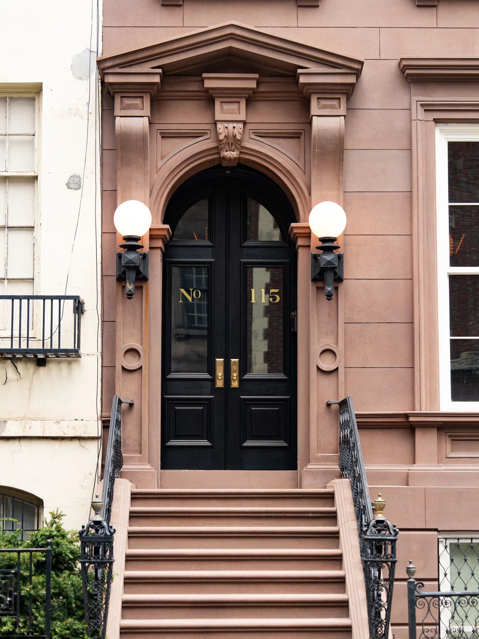 Carved warm-brown stone surrounds black double doors above a flight of steps, with ornate black iron railings and two globe lights.