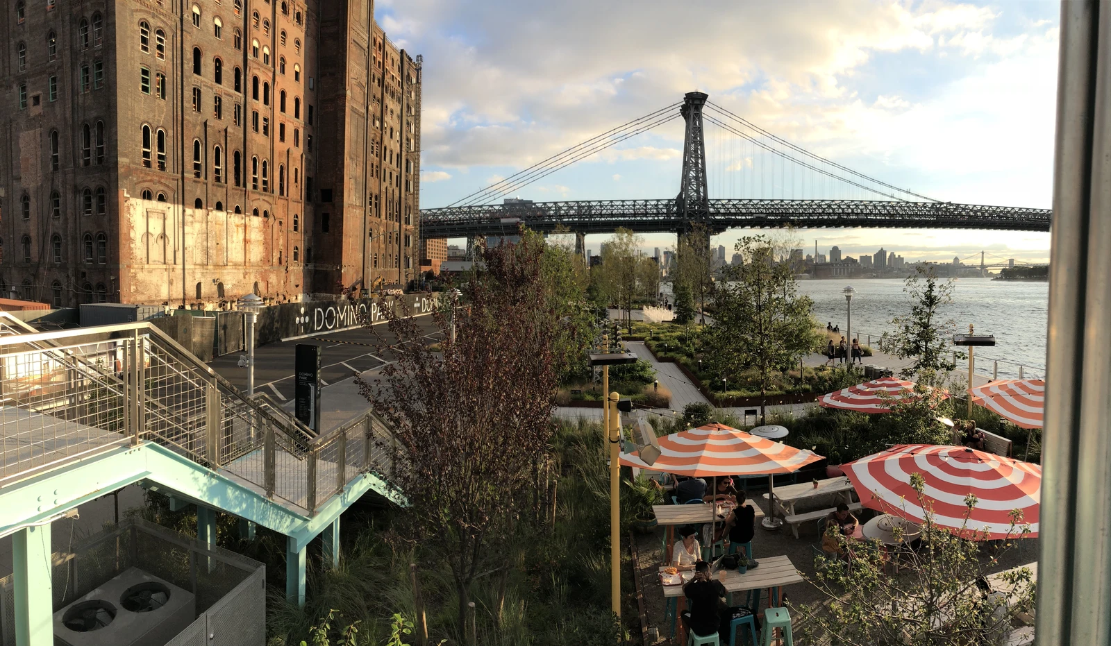 Warm evening light across the former Domino refinery, a turquoise elevated walkway, planting and orange-striped umbrellas beside the East River and Williamsburg Bridge.