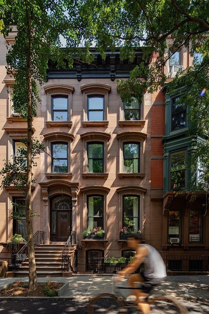 A cyclist passes a sunlit brownstone with tall windows, an iron-railed stoop and leafy trees.