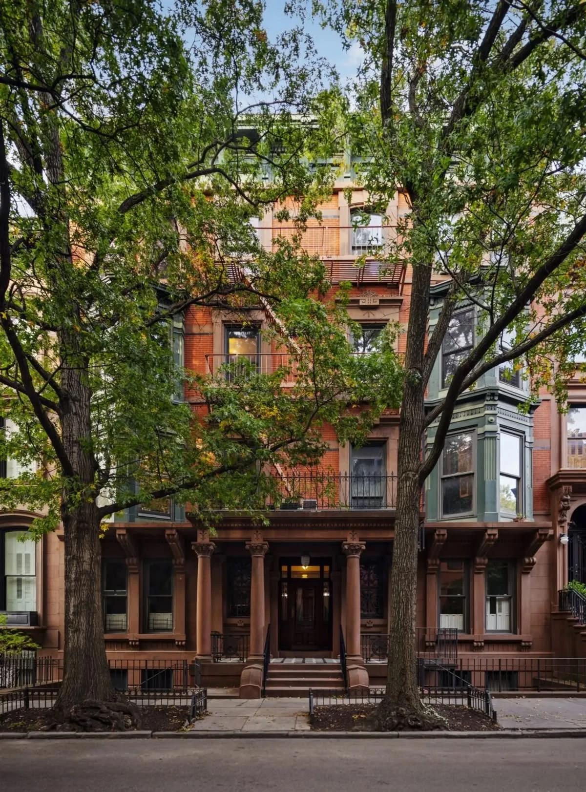 A red-brick and brown-stone townhouse with a columned entrance and bay windows, framed by leafy trees.