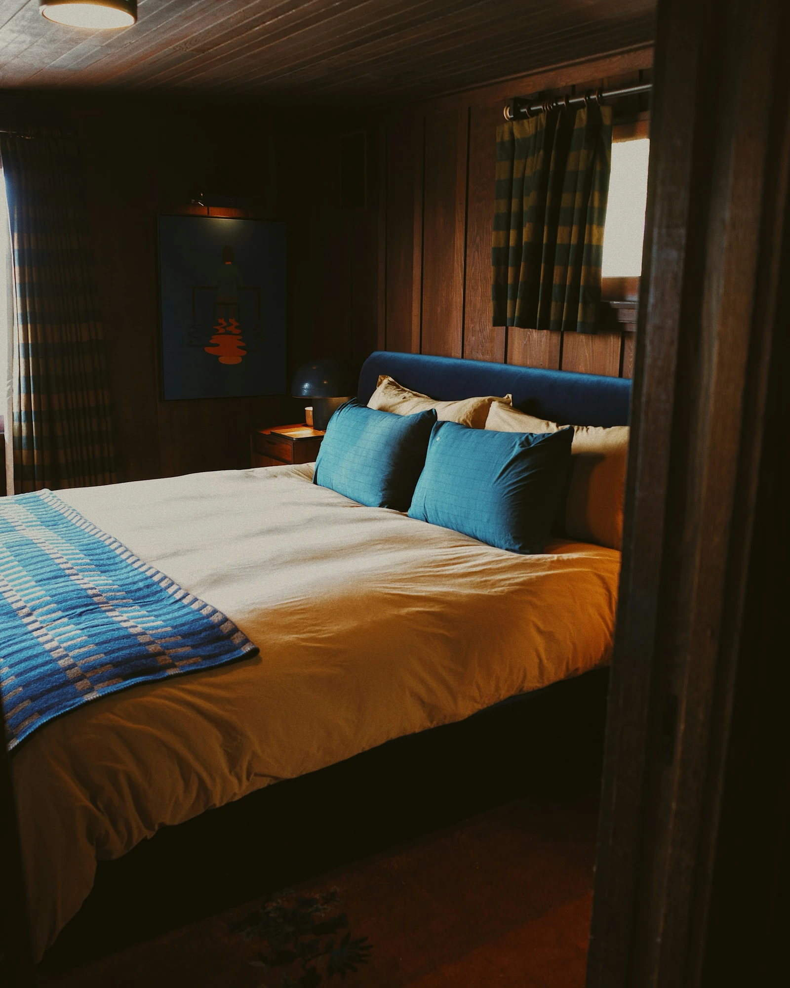A bedroom with cream and amber bedding, blue cushions, dark timber paneling and striped curtains.