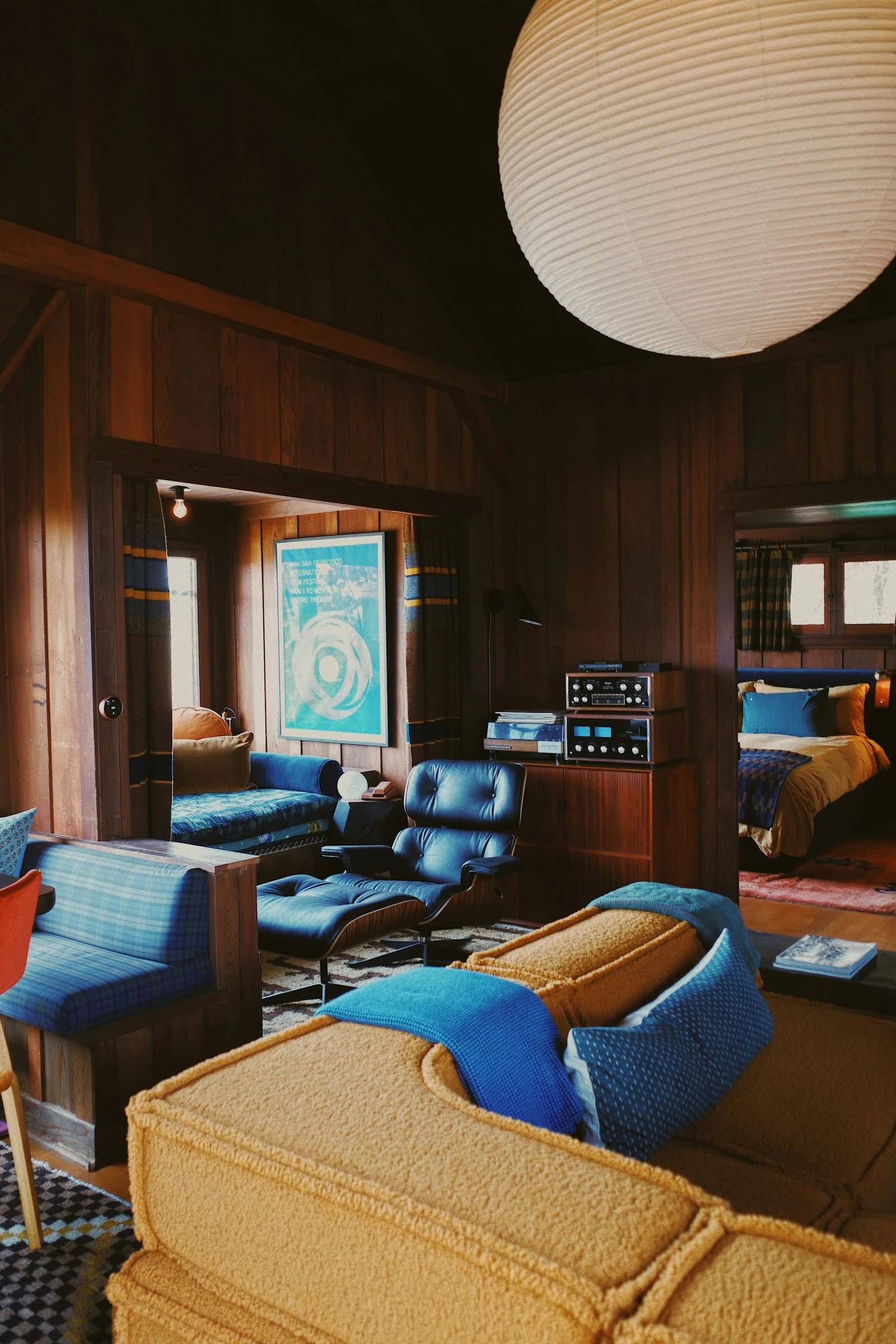 Mustard sofa and blue lounge chair beneath a large white paper lantern in a timber-paneled Laguna Beach cabin.
