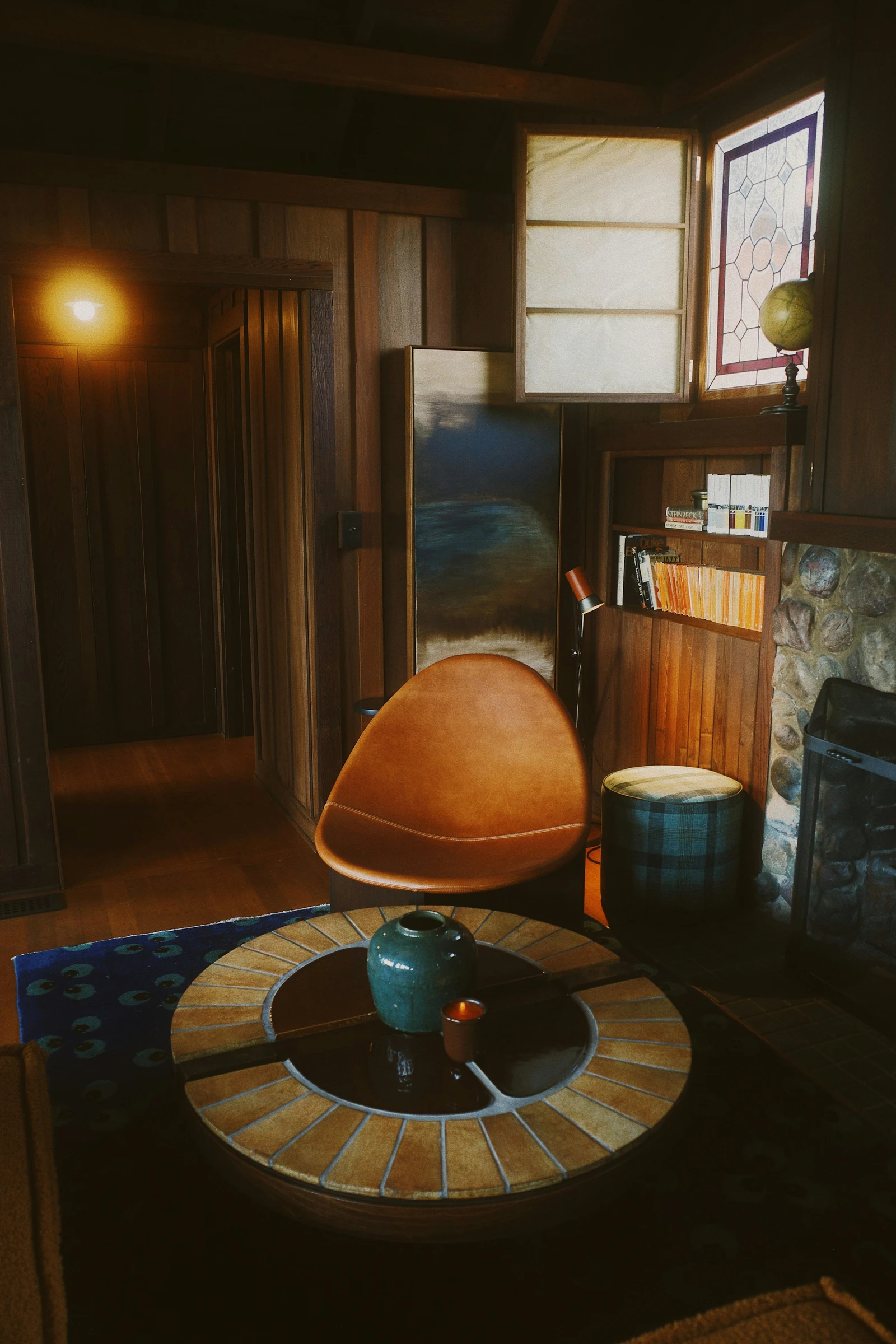 Amber armchair beside a stone fireplace, with wood-paneled walls, decorative glass and a round tiled coffee table.