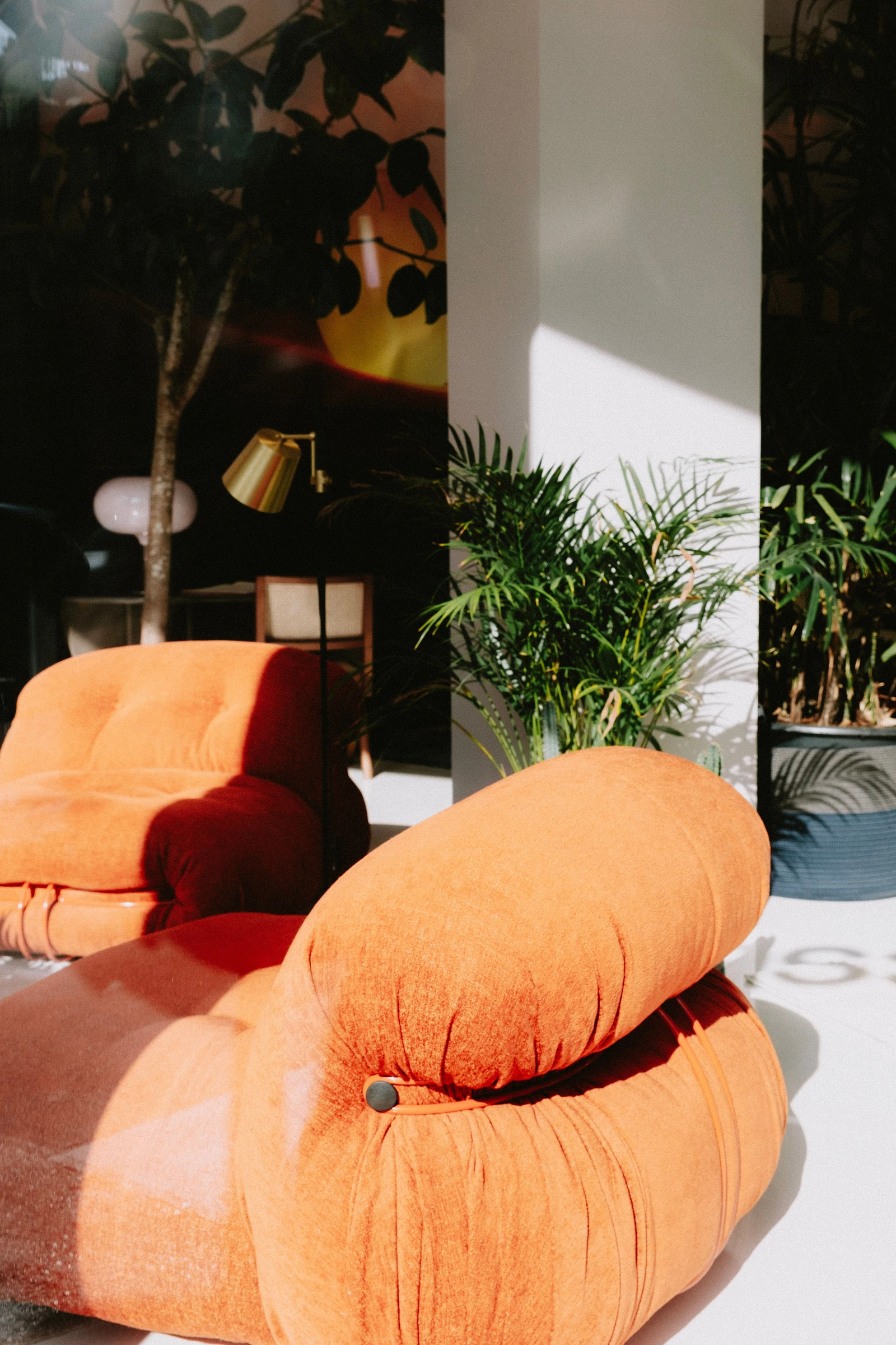Two rounded orange upholstered chairs beside a pale column, with dark circular fittings, indoor foliage and strong sunlight.