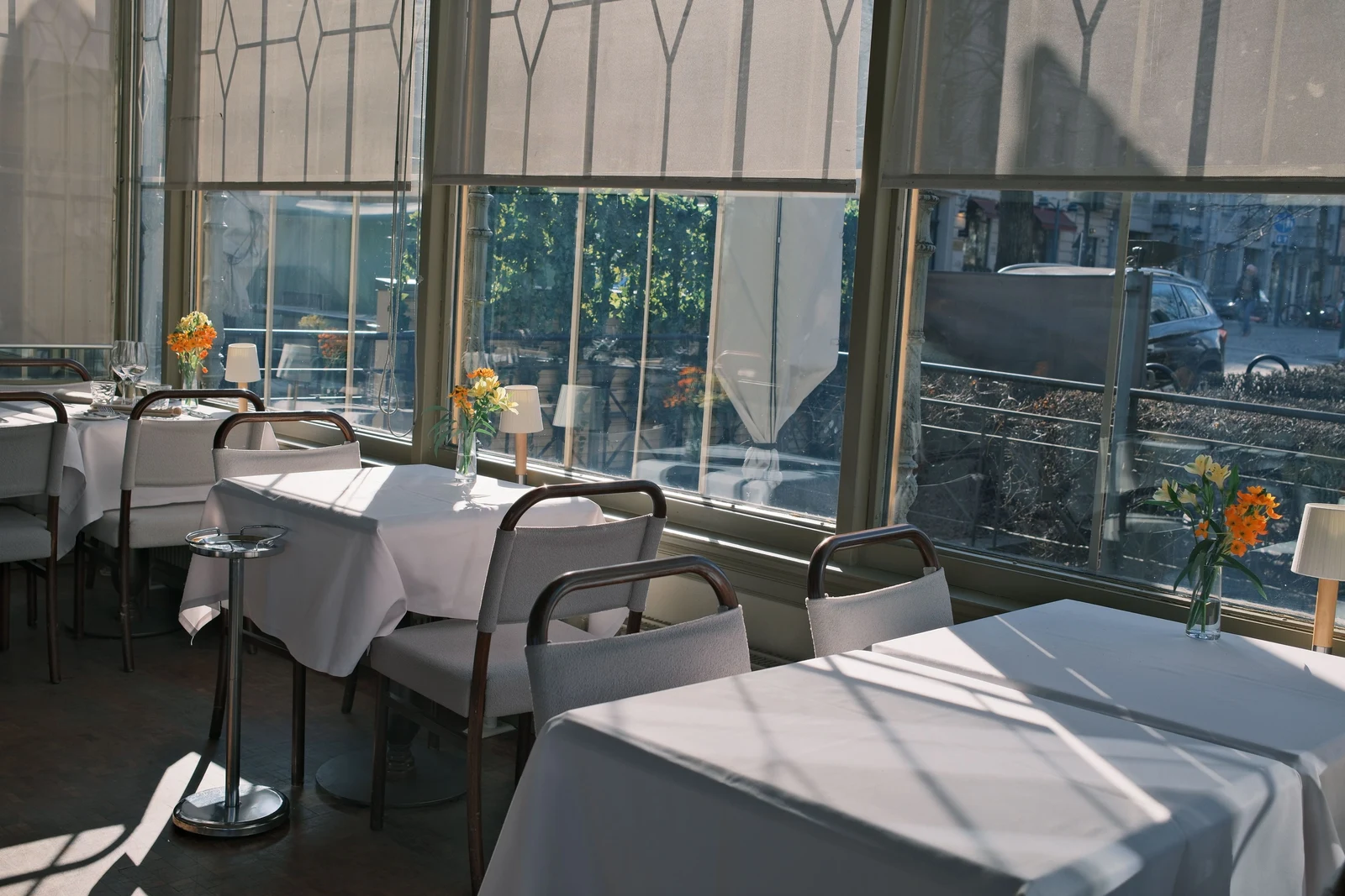 Sunlight falls across white-clothed restaurant tables beside tall windows, with small vases of yellow and orange flowers.